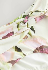 Gros plan sur un tissu blanc imprimé de motifs floraux roses et verts, torsadé au centre, montrant une texture lisse et des plis doux.