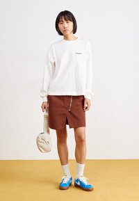 Camisa blanca de manga larga con logotipo, falda de mezclilla marrón con cremallera frontal, zapatillas azules y bolsa beige, todo frente a un fondo amarillo.