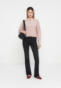 Ljusrosa kort cardigan med knäppning, kombinerad med svarta utsvängda jeans. Modellen håller en svart axelväska och har på sig röda skor.
