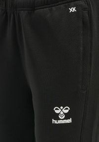 Melnas sporta bikses, kas izgatavotas no gludas auduma, ar elastīgu jostu, sānu kabatām un baltu logotipu kreisajā kājā.