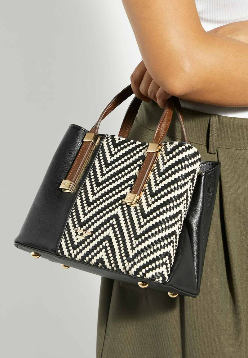 Femme tenant un sac à main tissé en noir et blanc avec un motif en zigzag, des poignées marron et des accents dorés, portée sur un pantalon vert olive et un haut blanc.