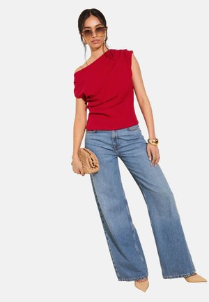 Femeie purtând un top roșu cu un umăr gol, blugi albaștri deschiși cu pantaloni evazați, pantofi bej cu vârf ascuțit, ținând un clutch bej țesut și purtând ochelari de soare și o brățară aurie.
