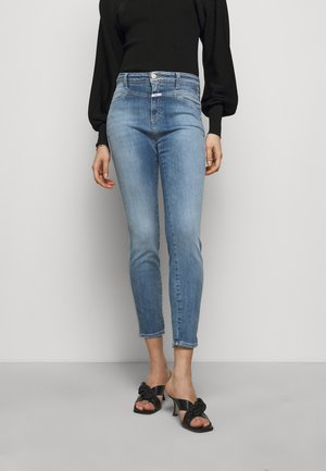 Femme portant un jean skinny taille haute bleu clair, un haut noir à manches bouffantes et des sandales à talons noires avec un détail en nœud, se tenant sur un sol gris.