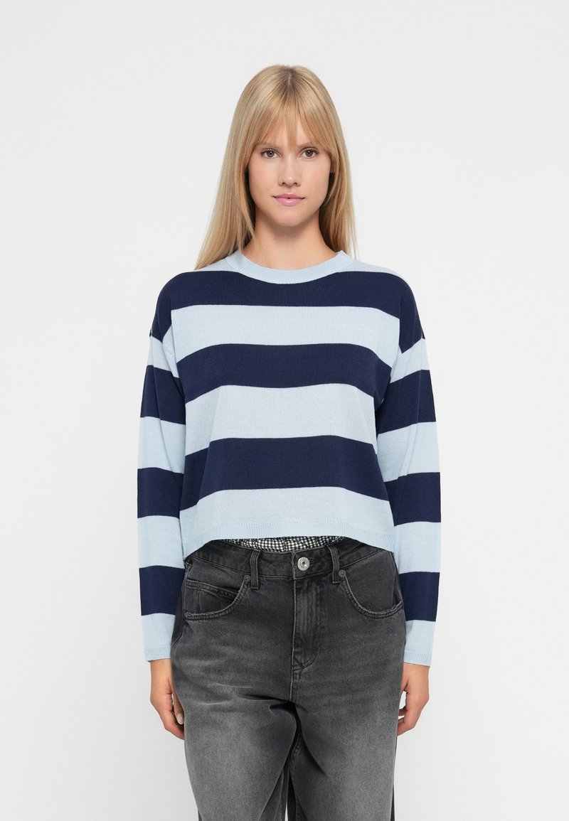 Woman wearing a blue and navy wide-striped sweater with long sleeves and high-waisted faded black jeans against a white background.