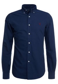 Námořnická modrá košile s dlouhým rukávem, s límečkem na knoflíky, bílými knoflíky a malým červeným logem na hrudi. Vyrobena z bavlny. Hladký povrch.