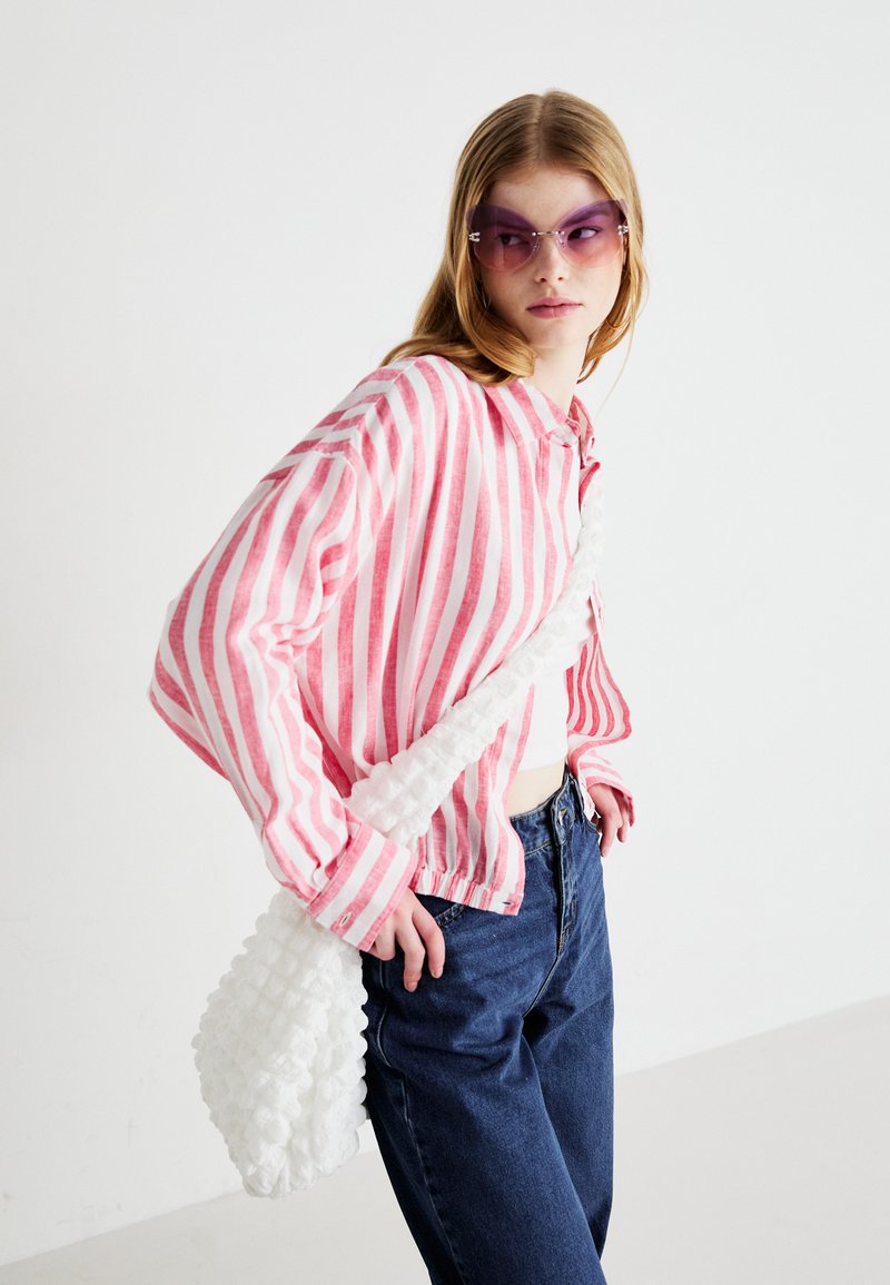 Różowo-biały w paski koszula z guzikami, luźny krój, w zestawieniu z ciemnoniebieskimi dżinsami o wysokim stanie. Biała torebka o fakturze na ramieniu i fioletowe okulary przeciwsłoneczne.