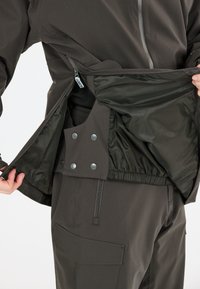 Mørk jakke med lynl�åsåbning, der har et glat indre for og tryknapper i taljen. Stoffet ser vandafvisende og holdbart ud.