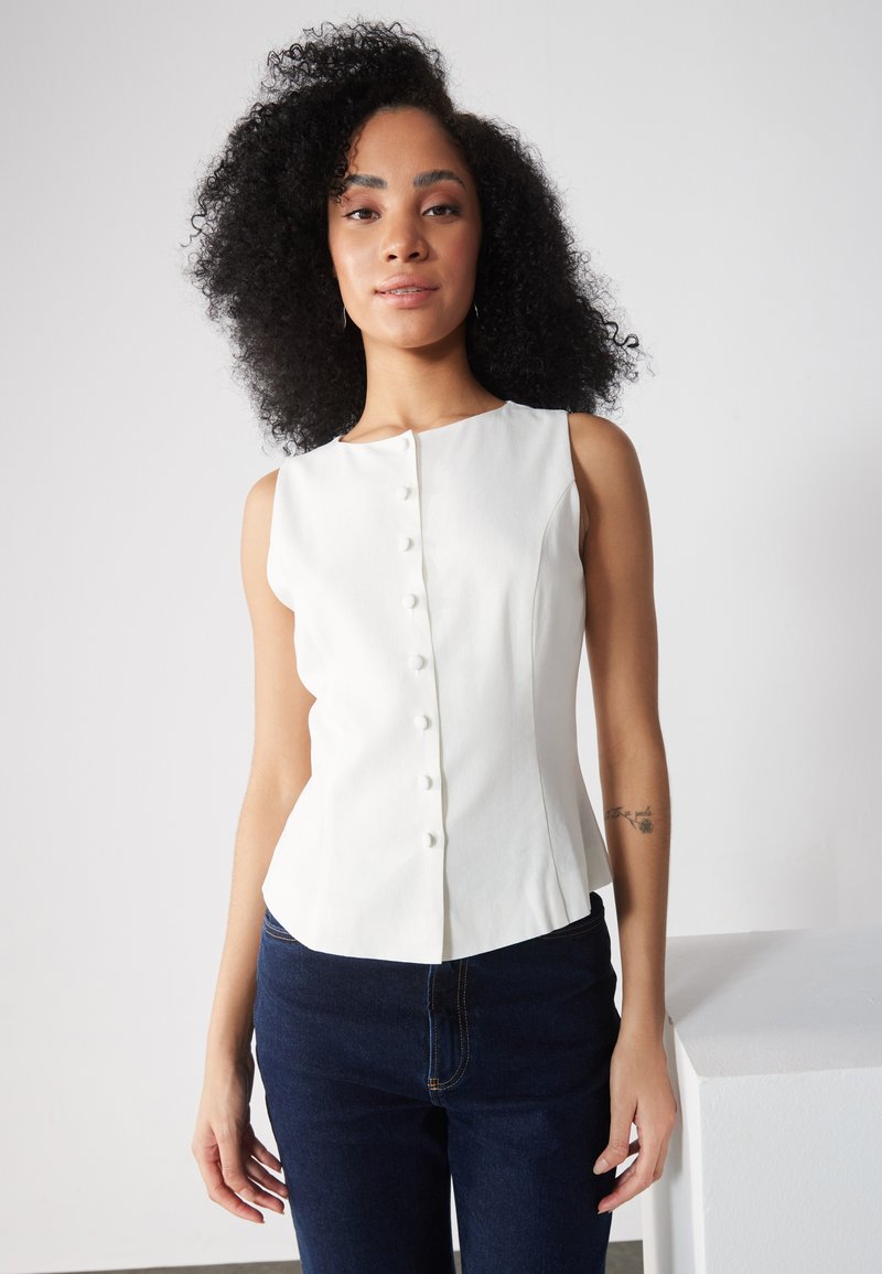 Hvid ærmeløs bluse med høj hals, der har en frontlukning med knapper og glat stof. Sammen med mørkeblå jeans.