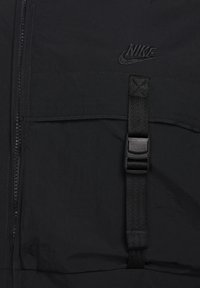 Černá bunda Nike s předním zipem, texturovanou tkaninou, nastavitelným páskem s přezkou a vyšitým logem s matným povrchem.