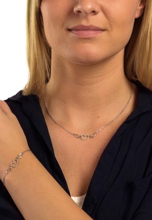 Collier et bracelet en argent présentant des formes ovales et des cœurs entrelacés. La chaîne a une finition polie avec une texture visible.