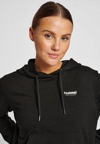 Svart hoodie i bomullsblandning, med justerbara dragband, en frontficka och vit "hummel"-logotyp på vänster bröst.