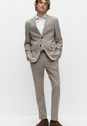 Jonge man staand met handen in de zakken, gekleed in een lichtgrijs pak, wit overhemd, bruine riem en bruine nette schoenen tegen een effen achtergrond.