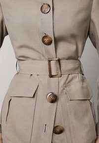 Beżowa kurtka z paskiem, z dużymi brązowymi guzikami, dwoma przednimi kieszeniami i gładką teksturą. Posiada detale w postaci przeszyć oraz zdefiniowaną sylwetkę.