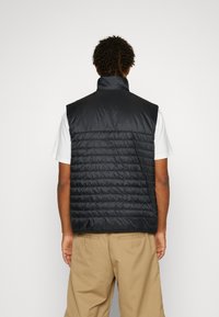 Svart pufferväst med hög krage, quiltad textur och ärmlös design, tillsammans med en vit T-shirt och beige byxor.