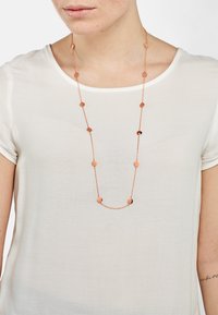 Collana lunga in oro rosa con anelli di forma circolare e ovale, catena testurizzata, indossata sopra una maglietta bianca. Design semplice, materiale leggero.