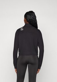 Top sportivo a maniche lunghe nero con colletto, caratterizzato da un logo sportivo sulla spalla e un design aderente, abbinato a leggings neri.