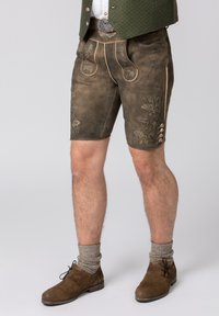 Lederhosen en cuir brun avec des coutures décoratives et des poches, ornés de boutons en laiton sur le côté. Portés avec des chaussettes jusqu'aux genoux grises et des chaussures brunes.