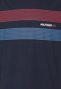 Camiseta azul marino con rayas horizontales en rojo y azul claro. Tiene el texto "HILFIGER '85" en blanco. Fabricada con una tela suave y transpirable.