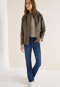 Femme debout, main dans la poche, portant une veste à capuche marron, un pull beige, un jean bleu et des baskets argentées, sur un fond neutre.