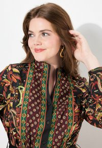Camicia nera con vivaci motivi floreali e geometrici in rosso, verde e giallo. Presenta un alto colletto e chiusura con bottoni.