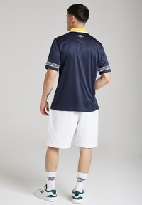 Cămașă sport de culoare bleumarin cu guler galben, manșete cu model și pantaloni scurți albi. Purtați cu adidași albi și șosete cu model, privit din spate.