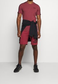 Camiseta deportiva en color burdeos con detalles en negro, combinada con pantalones cortos burdeos que presentan un gran logo negro. Suéter oscuro atado alrededor de la cintura. Zapatillas negras.
