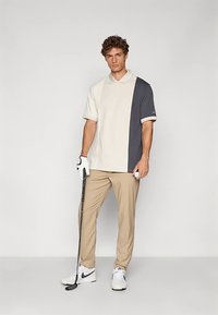 Pikétröja med ett blockdesign i kräm och mörkgrå färg, korta ärmar och krage. Beiga byxor och vita golfsandaler kompletterar outfiten.