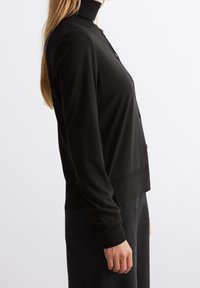 Schwarze Langarm-Cardigan aus weichem Stoff, mit hohem Kragen, durchgehender Knopfleiste und geripptem Saum. Glatte Textur und figurbetontes Design.
