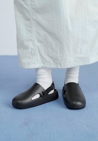 Sorte gummiklær med utskårne detaljer, justerbar stropp og teksturert såle, iført med hvite sokker og et lys blått plagg.