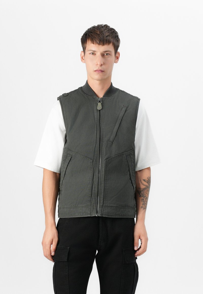 Chaleco sin mangas de color verde oscuro con cremallera frontal, dos bolsillos laterales y un pequeño bolsillo frontal. Lleva puesto una camiseta blanca de manga corta.