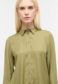 Camicia verde oliva con bottoni, dal tessuto liscio, maniche lunghe e colletto classico. Presenta bottoni abbinati lungo la patta frontale.