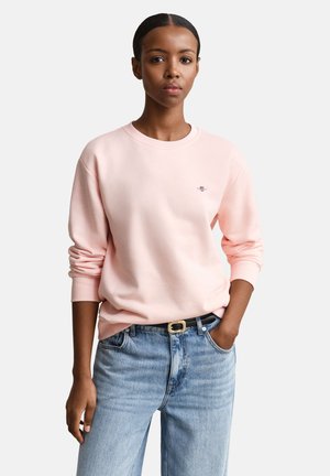 Kvinne iført lys rosa sweatshirt og lyseblå jeans, stående med en hånd i lommen mot ensfarget bakgrunn.