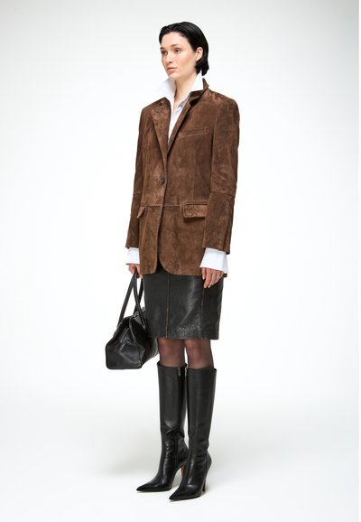 Nő barna szarvasbőr blézerben, fehér ingben hosszított mandzsettákkal, fekete bőrszoknyában, fekete térdzokni csizmában, és fekete bőr táskával a kezében.