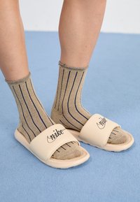 Sandales en toile beiges avec une large sangle et un logo noir, portées avec des chaussettes rayées dorées sur une surface bleue texturée.