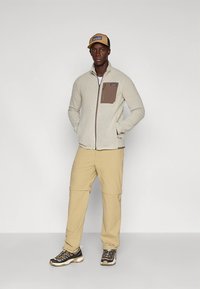 Kreemika värvi fliisjope pruunide rinnataskutega, kombineeritud beežide pükste ja mustade matkajalatsitega, ühevärvilise taustaga.