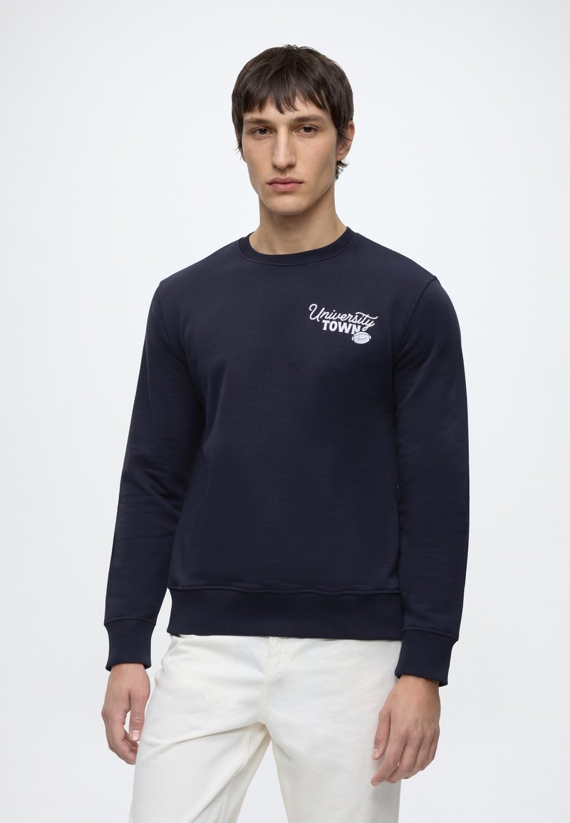 Ung mand iført en mørkeblå sweatshirt med teksten "University Town" og hvide bukser, stående mod en ensfarvet lys baggrund.