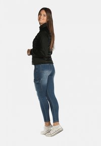 Veste rembourrée noire, leggings en denim bleu avec poche cargo et baskets blanches. Textures lisses et design ajusté. Pose debout.