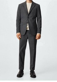 Homme portant un costume gris foncé sur mesure avec un pull en maille blanc et des bottes en cuir noires, debout devant un fond uni.