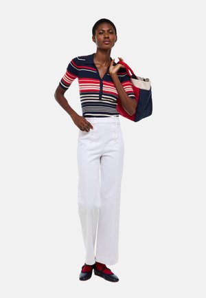 Femme portant un t-shirt à manches courtes rayé bleu marine, rouge et blanc, un pantalon blanc taille haute, des chaussettes rouges, des ballerines bleu marine, tenant un sac fourre-tout rouge et bleu marine.
