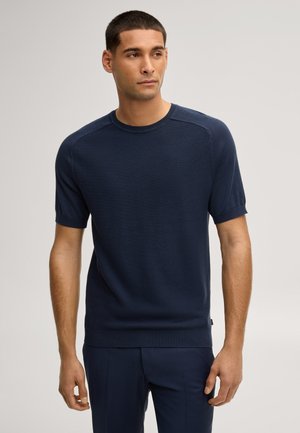 Uomo con capelli corti che indossa una maglia a maniche corte in maglia testurizzata blu navy e pantaloni blu navy, che guarda leggermente di lato.