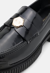 Zwarte gepatineerde leren loafer met een gouden hexagonaal logo op de band, voorzien van textuurdetails en een geribbelde rubberen zool.