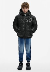 Sort dunjakke med hætte, dekoreret med strukturerede edderkoppespind- og karakterdesigns. Kombineret med blå denimjeans og sorte sneakers.