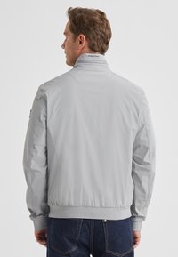 Hellgraue Bomberjacke mit Stehkragen, gerippten Bündchen und Saum. Glatte Textur, minimalistisches Design und dezentes Logodetail am Kragen.