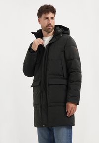 Man in een zwarte winterjas met capuchon en grote voorzakken, in blauwe spijkerbroek, die de kraag van de jas aanpast, tegen een plain witte achtergrond.