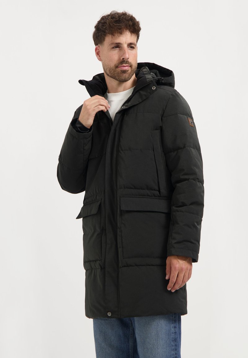 Man in een zwarte winterjas met capuchon en grote voorzakken, in blauwe spijkerbroek, die de kraag van de jas aanpast, tegen een plain witte achtergrond.