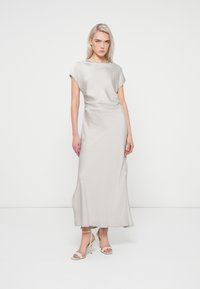 Robe longue, gris clair, avec une texture lisse, des manches courtes et un décolleté arrondi. Associée à des sandales à talons nus.