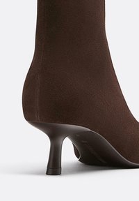 Ruskea mokkanahkainen nilkkuri, jossa on kapeakärkinen piikkikorko, sileä pinta ja tyylikäs, vartalonmyötäinen muotoilu.