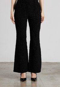 Pantalones de campana negros fabricados en un tejido texturizado, con un patrón intrincado. La modelo lleva tacones negros de punta.