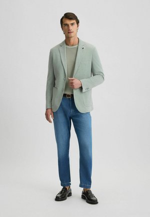 Blazer verde chiaro con due bottoni, revers a punta e tasche. Abbinato a una camicia grigia chiara e jeans blu. Mocassini neri completano il look.