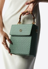 Zielona, pleciona torba o wyraźnym kształcie, z bambusowym uchwytem, złotymi detalami i zapięciem na klapkę z logo. Widoczne detale materiału teksturowego.